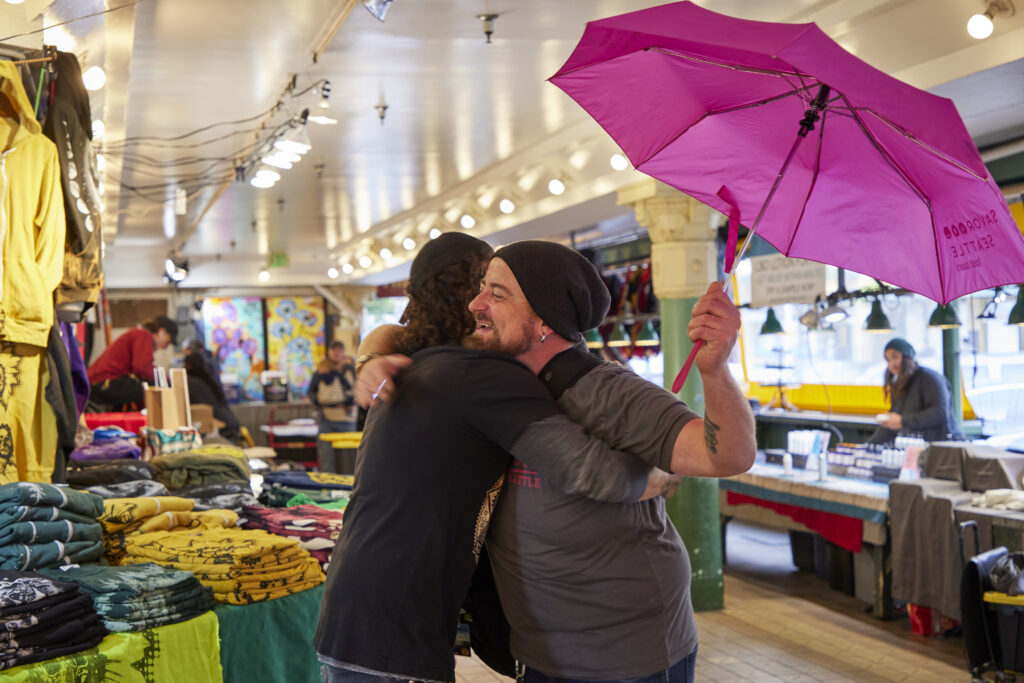 Tour guide Matthew Bentley hugs a vendor market while conducting a food tour of pike place market. He is holding up the iconic pink umbrella of Savor Seattle Tours