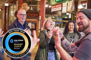 People on a food tour in Pike Place Market laugh about something the tour guide has said. Includes a logo that says that Savor Seattle Tours was voted one of the top ten food tours in the country